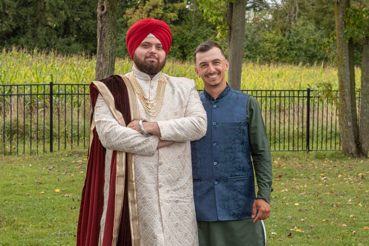 Two men in traditional South Asian attire stand outdoors smiling, one wearing a red turban and gold-embroidered sherwani.