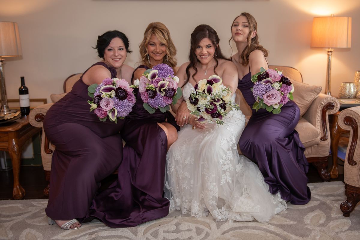 A bride in a white lace gown sits with three bridesmaids in purple gowns, all smiling and holding bouquets.