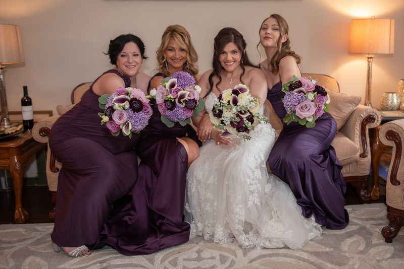 A bride in a white lace gown sits with three bridesmaids in purple gowns, all smiling and holding bouquets.