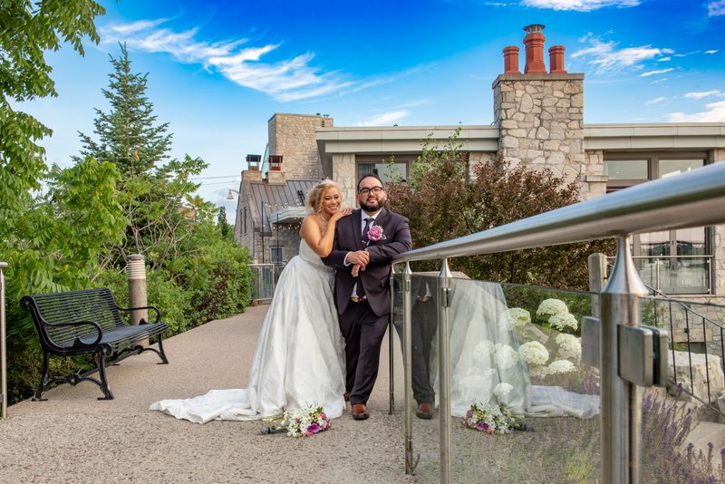 A smiling bride leans on her groom's shoulder on an outdoor terrace, surrounded by stone architecture.