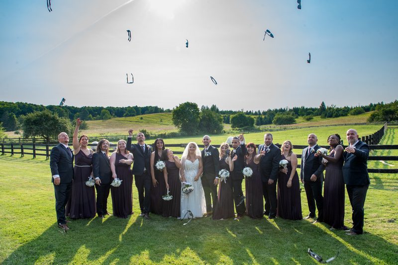 A wedding party in formal attire stands in a sunlit field as black and white confetti floats down around them.