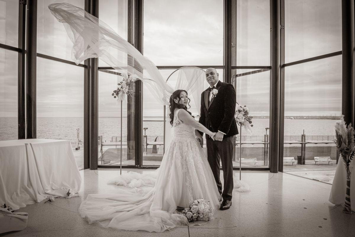 Bride and groom stand by windows overlooking waterfront, her veil billowing in wind, in soft monochrome light.