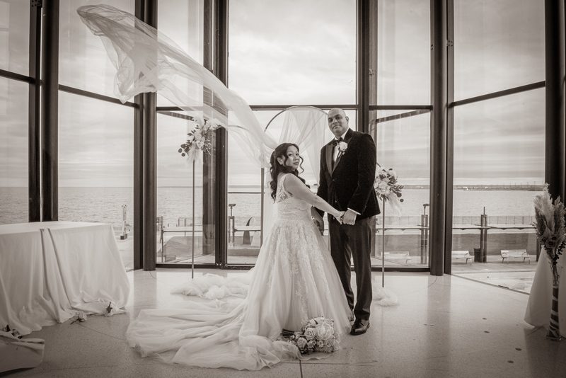 Bride and groom stand by windows overlooking waterfront, her veil billowing in wind, in soft monochrome light.
