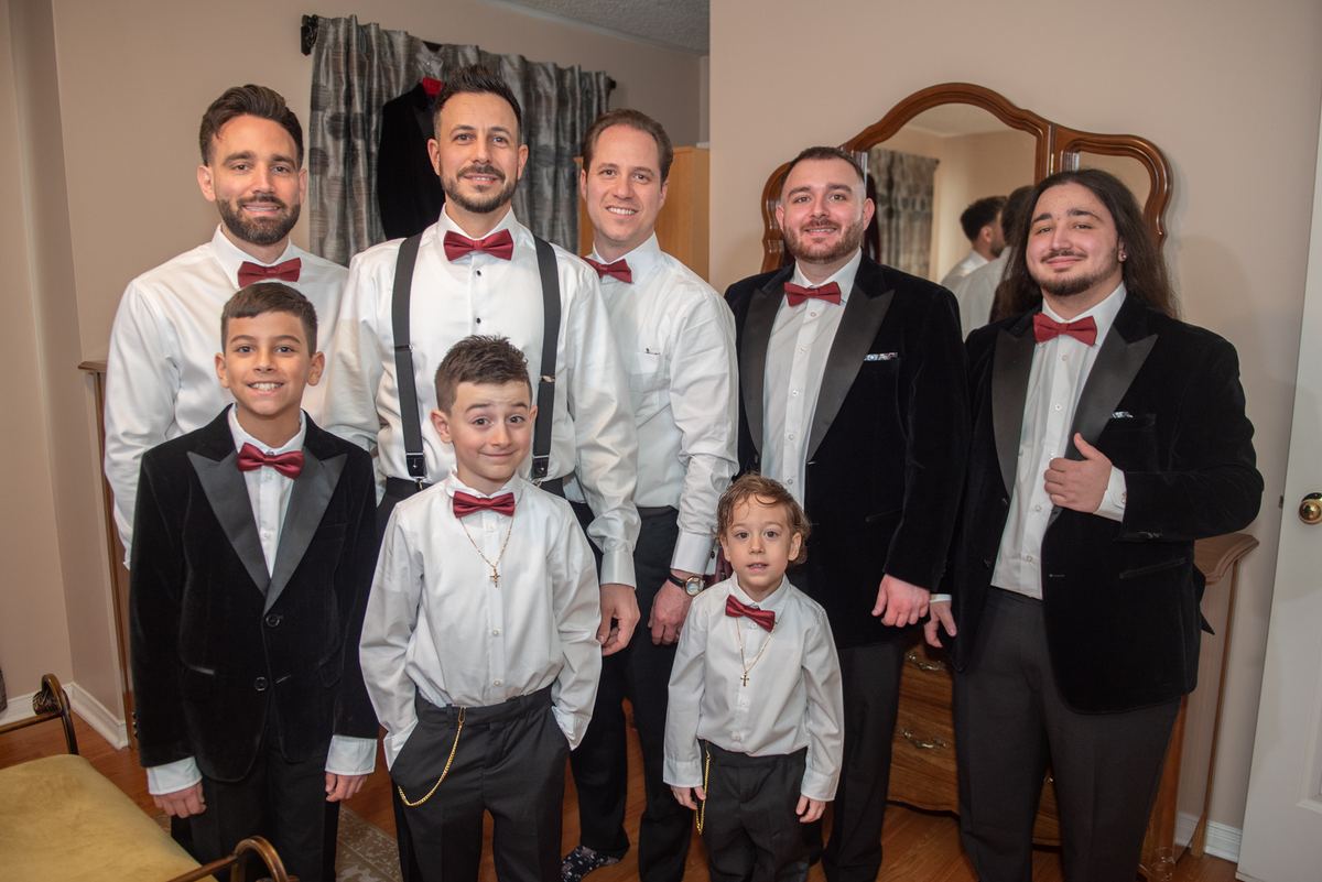 Men and boys in white shirts, dark trousers, red bow ties, some in velvet tuxedos, pose together indoors, smiling.