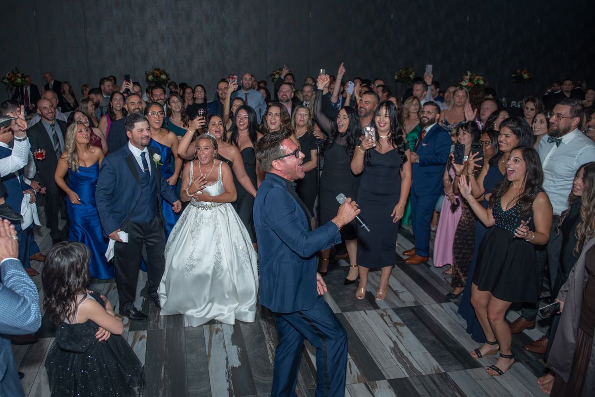 Bride and groom dance joyfully with guests cheering, clapping, and recording on phones at a lively reception.