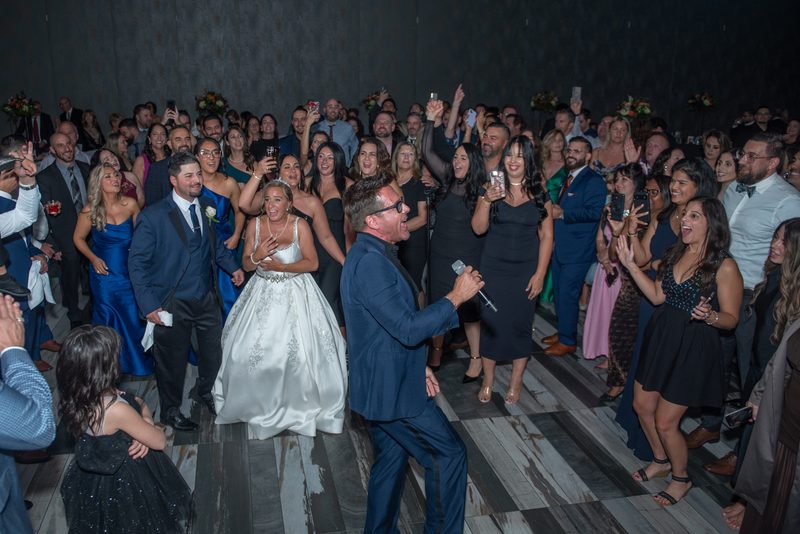 Bride and groom dance joyfully with guests cheering, clapping, and recording on phones at a lively reception.