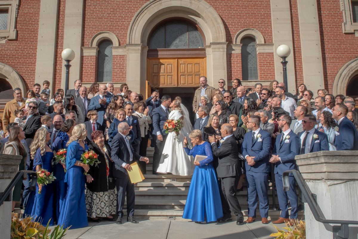Bride and groom exit brick church steps hand-in-hand, surrounded by cheering guests in formal attire.