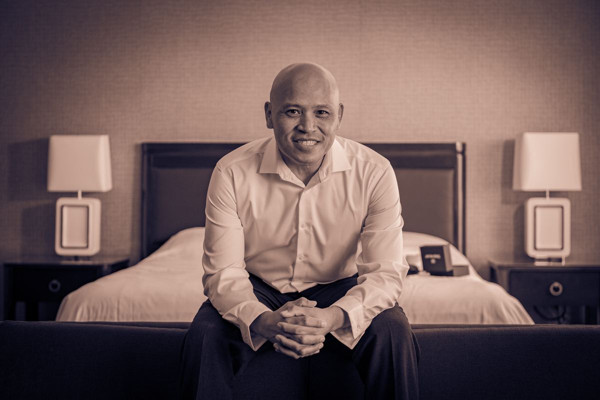 A bald man in a light-colored button-down shirt sits calmly on the edge of a dark sofa, smiling warmly at the camera.