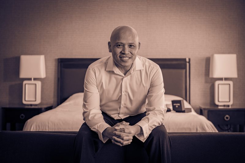 A bald man in a light-colored button-down shirt sits calmly on the edge of a dark sofa, smiling warmly at the camera.