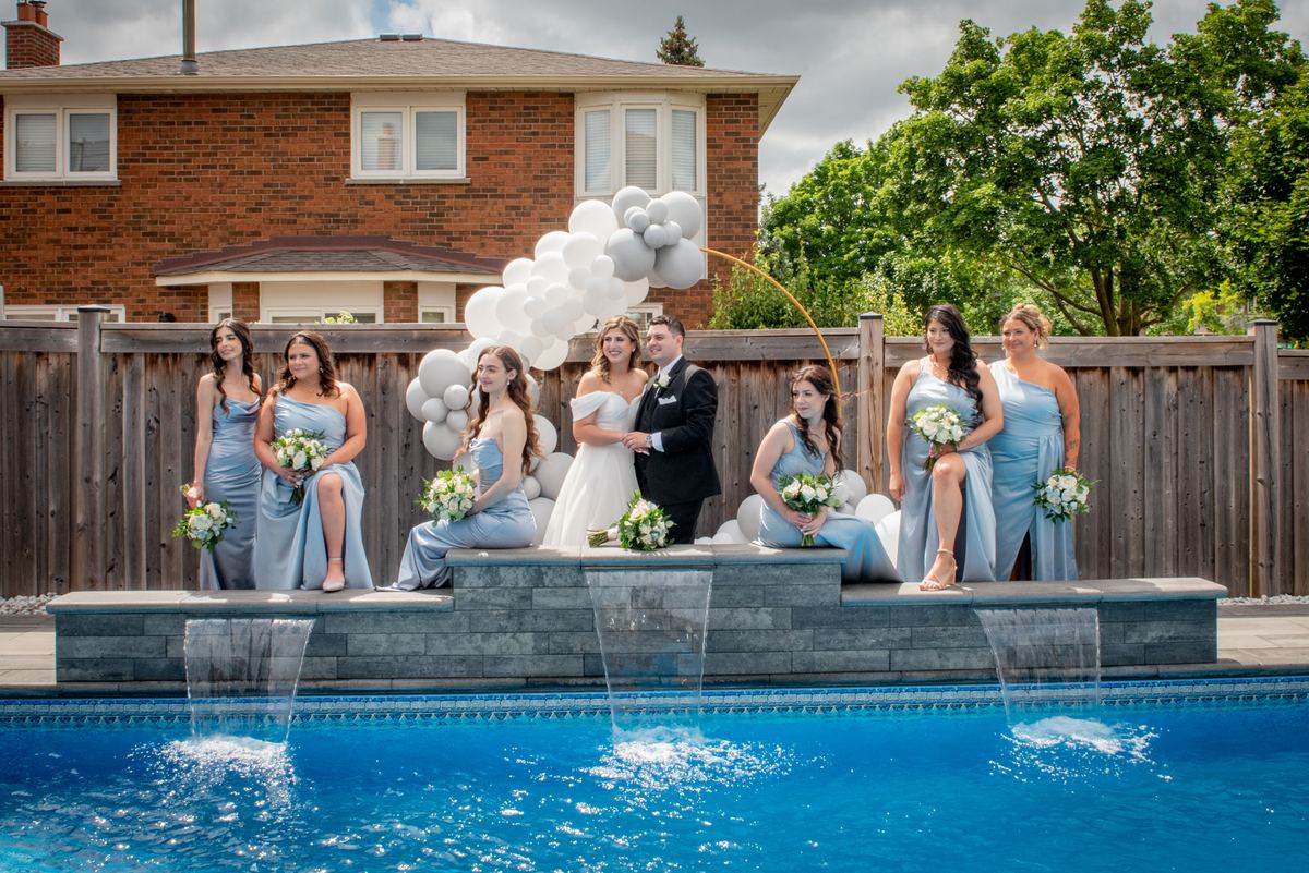 A bride and groom with bridesmaids pose by a pool with cascading water features, white balloons above them.