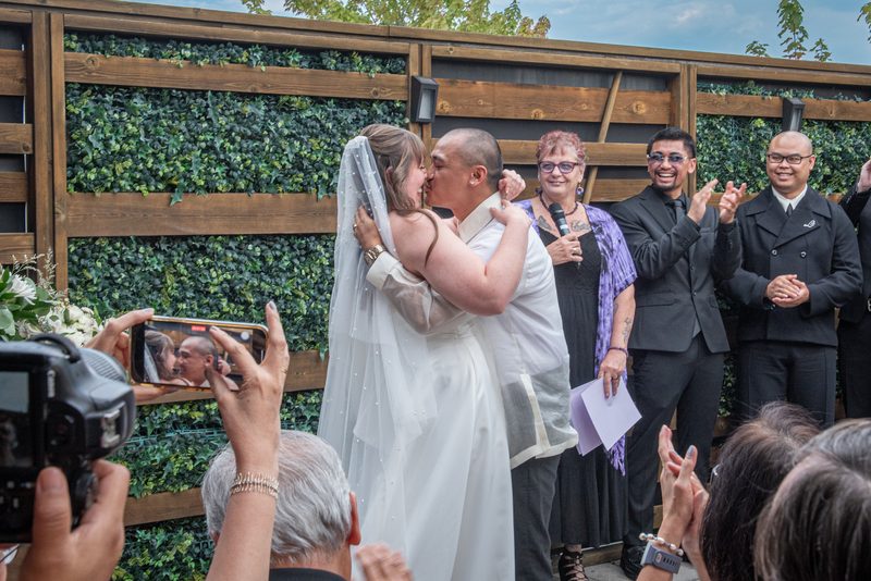 Bride and groom kiss joyfully among smiling guests and officiants near a green hedge, bathed in daylight.