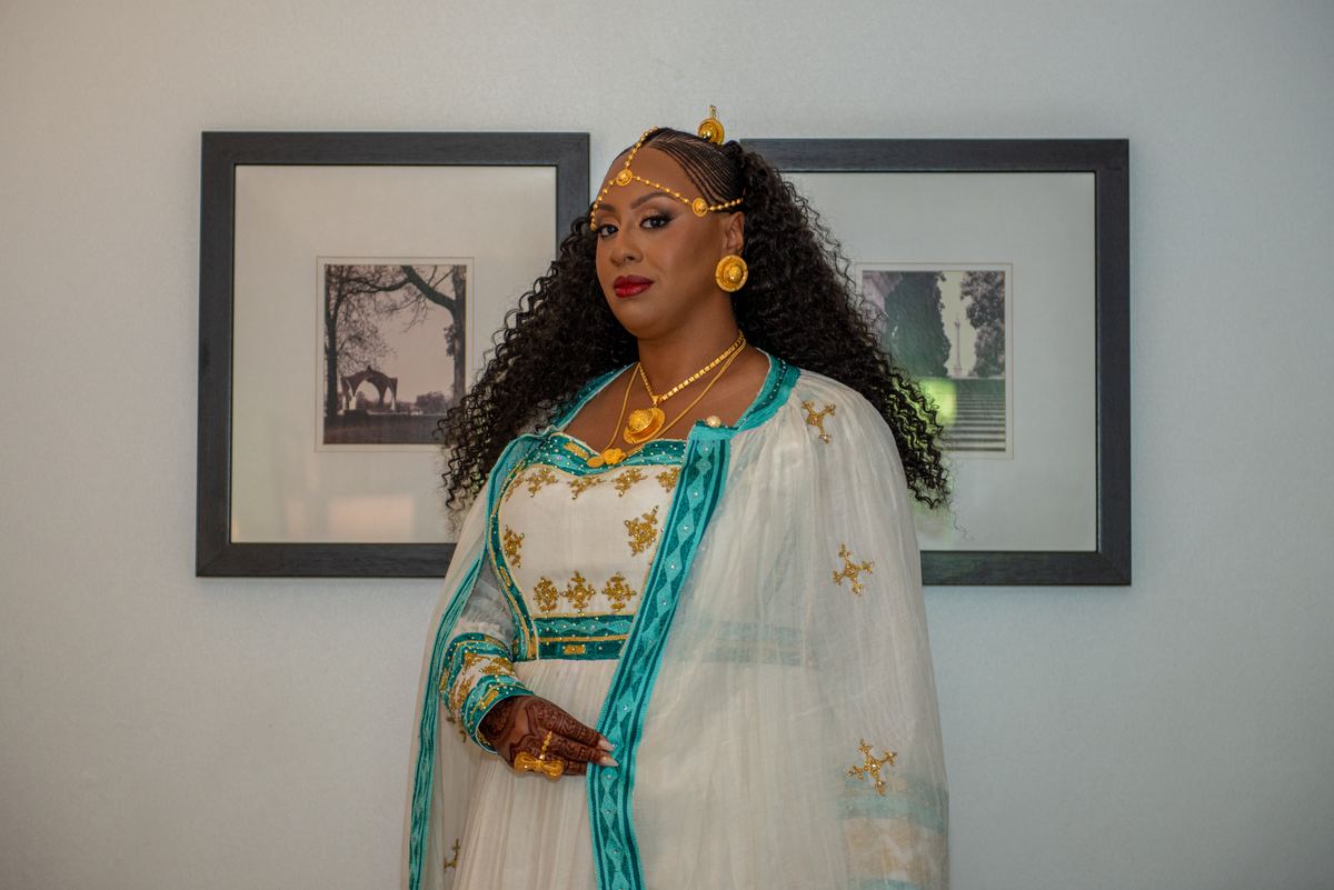 A woman in a white and teal traditional outfit with gold embroidery and jewelry stands calmly against a minimalist backdrop.