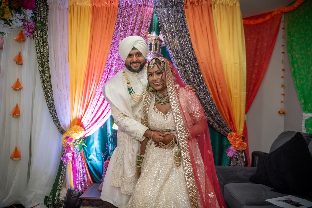 A Sikh bride in white and gold lehenga with red dupatta stands joyfully with her groom in ceremonial attire.