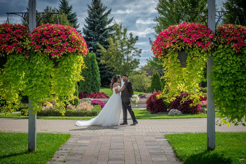 A bride and groom embrace along a garden path lined with pink hanging baskets under a partly cloudy sky.