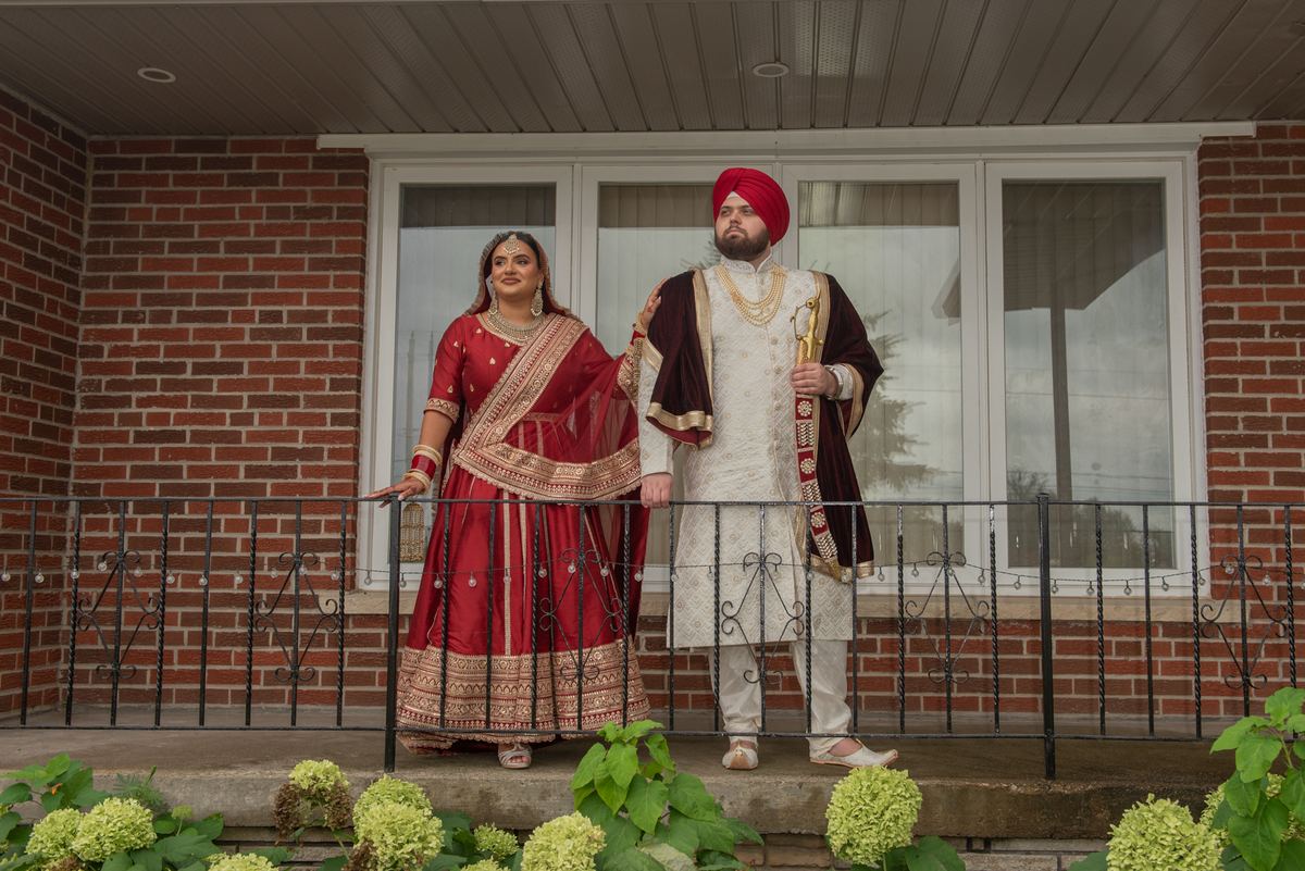 A bride in a red lehenga and a groom in a cream sherwani stand on a porch railing, posing for a photo.