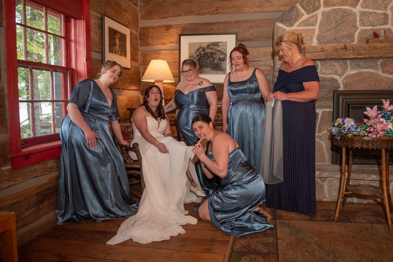 A bride in a white gown is seated among her laughing bridesmaids in blue satin gowns, illuminated by warm cabin light.