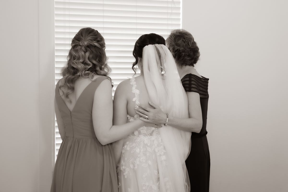 Three women in embrace by window, backs to camera, gazing out, diffused light filtering through blinds.