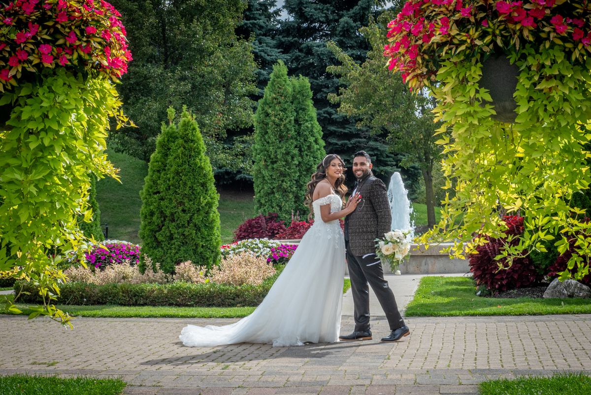 Bride and groom walk hand in hand on a brick path surrounded by lush greenery and pink hanging flowers.