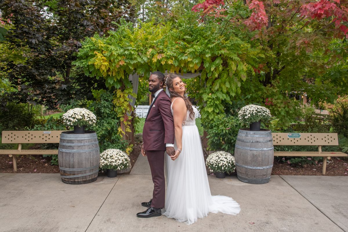 A smiling bride and groom in white and burgundy stand back-to-back in an autumn garden, framed by wooden benches.