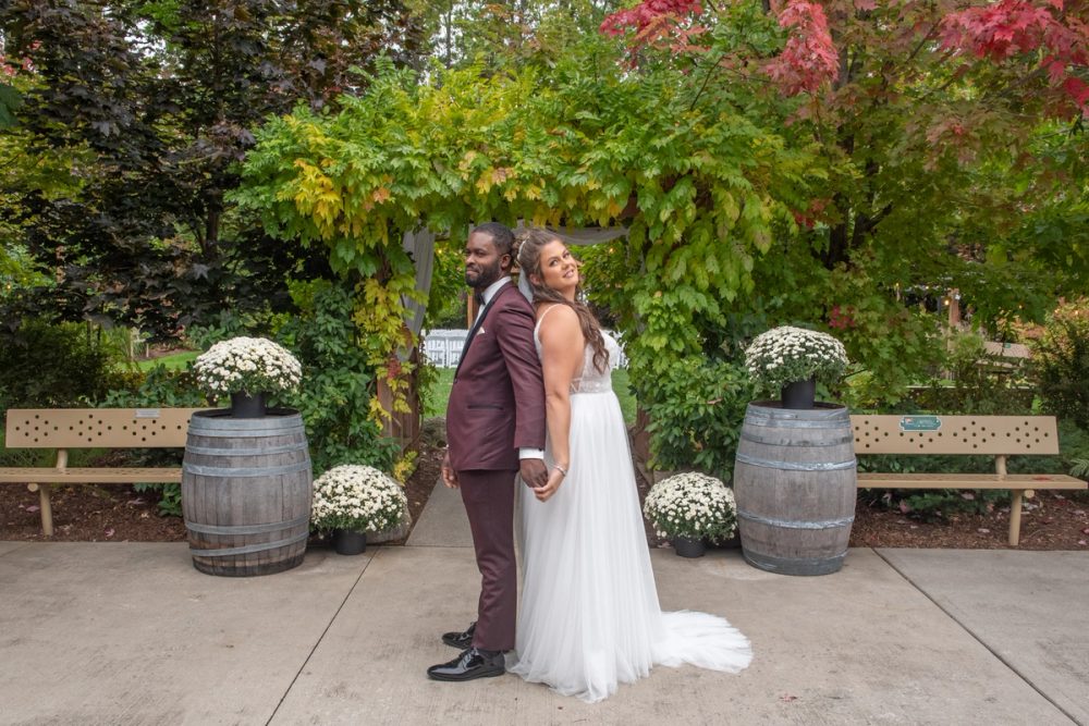 A smiling bride and groom in white and burgundy stand back-to-back in an autumn garden, framed by wooden benches.