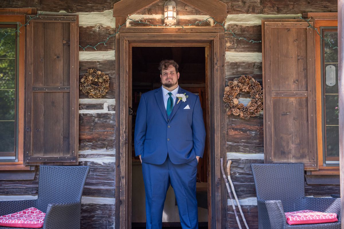 A groom in a blue suit stands by a rustic cabin door, hands in pockets, gazing at the camera, with pinecone wreaths.
