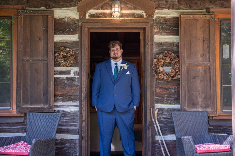 A groom in a blue suit stands by a rustic cabin door, hands in pockets, gazing at the camera, with pinecone wreaths.