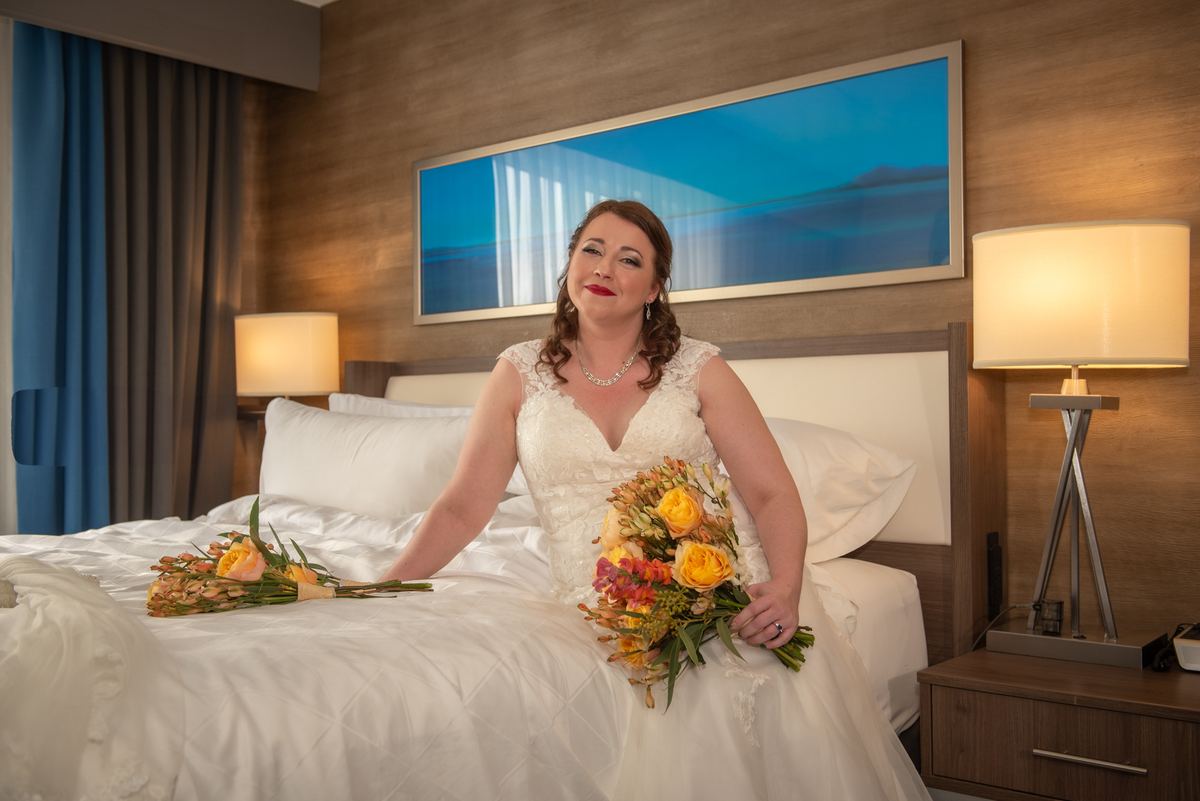 A bride in a white lace gown sits on a bed with flowers, smiling at the ceiling near artwork and lamps.