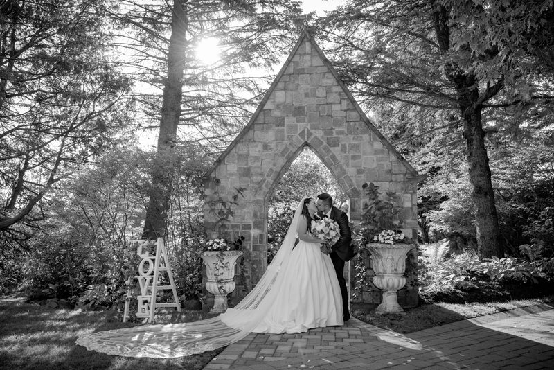 A bride and groom kiss beneath a stone Gothic archway in a sun-dappled garden, framed by trees and ornate planters.