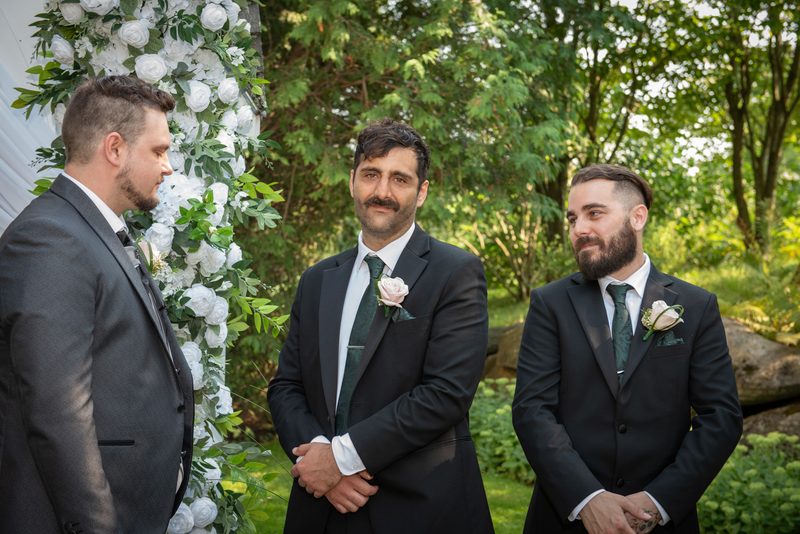 Three men in suits stand before a floral arch at an outdoor wedding, their expressions ranging from solemn to smiling.