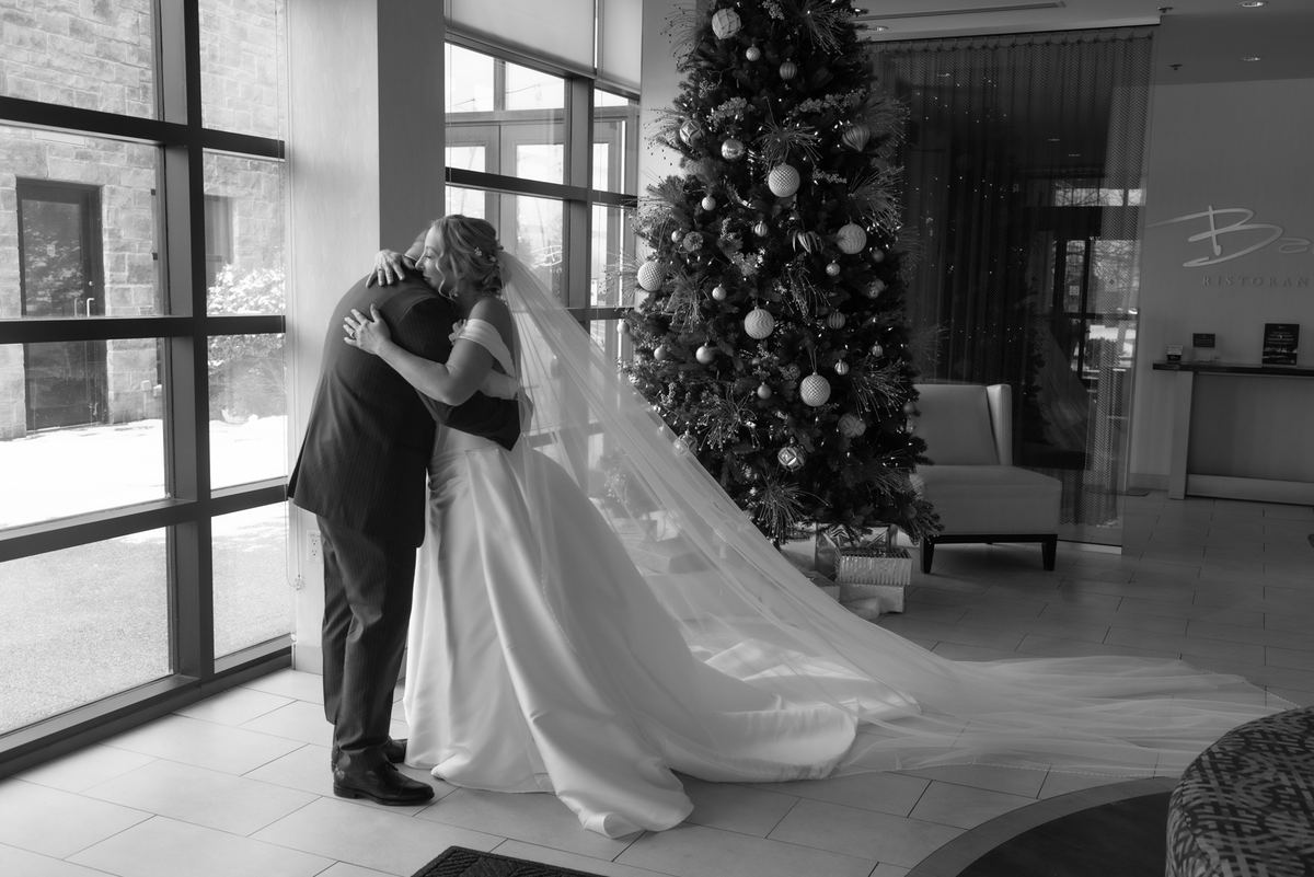A bride and groom embrace tenderly near a Christmas tree in a well-lit room with natural light streaming through windows.