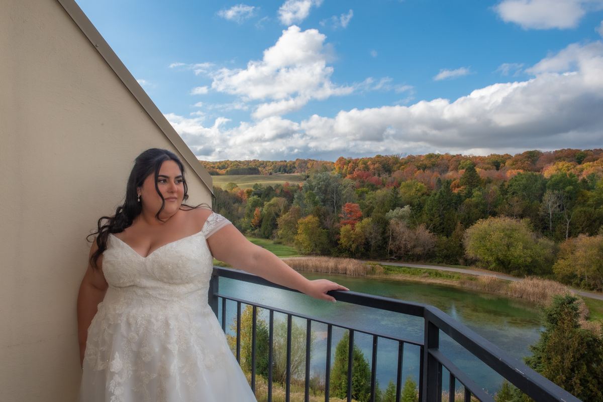 A bride in a white lace gown stands on a balcony overlooking an autumn landscape with colorful trees and a river.