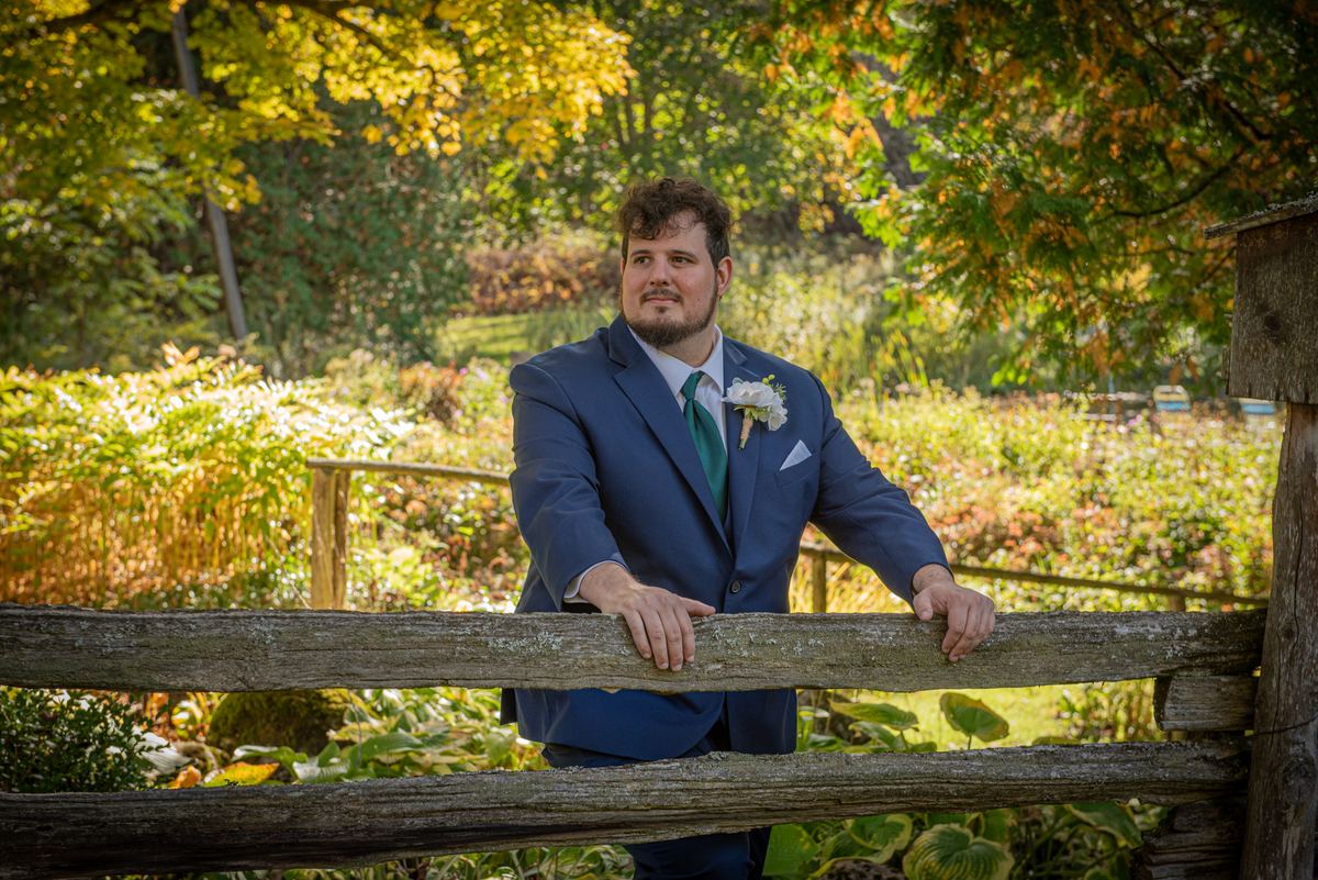 A groom in a navy suit and green tie leans against a rustic wooden fence, surrounded by autumn foliage and dappled sunlight.