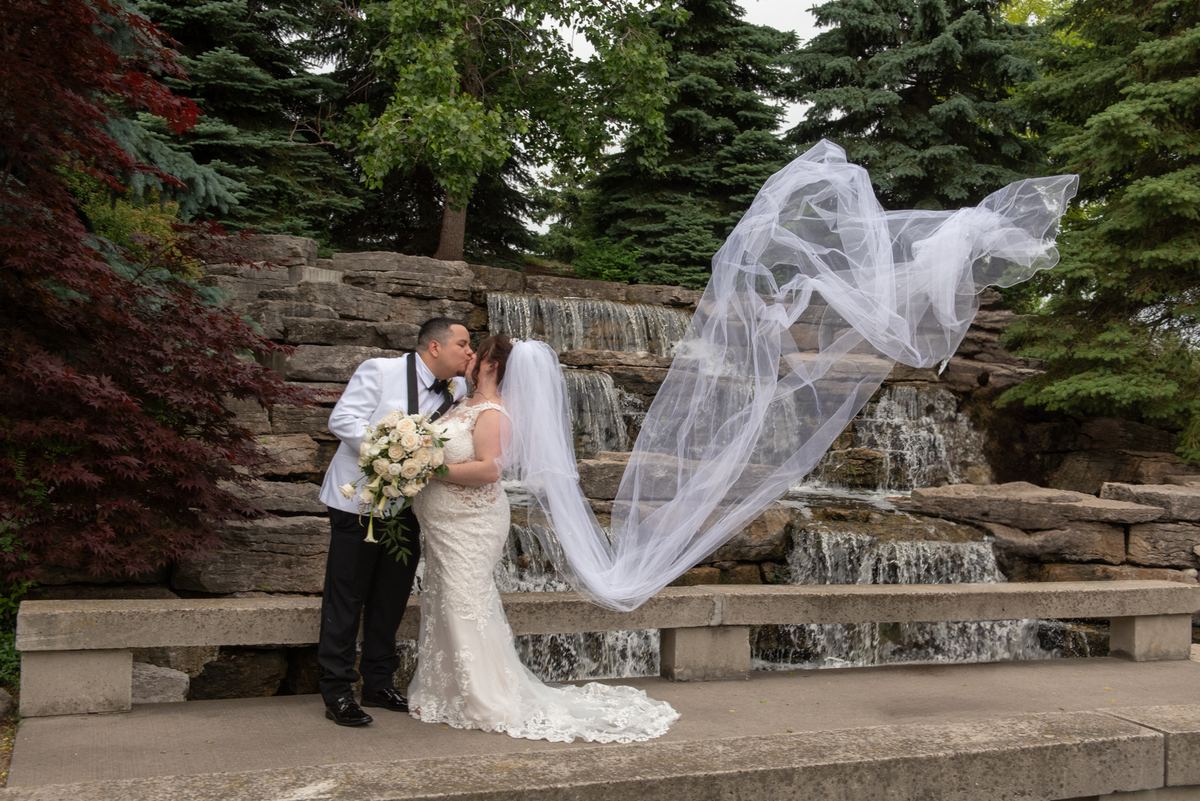 A bride and groom kiss on a stone bench, surrounded by lush greenery and a cascading rock waterfall.