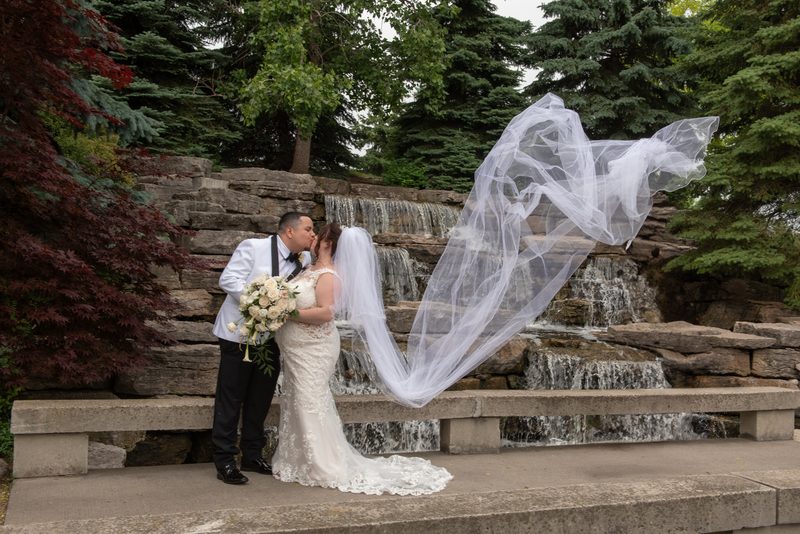 A bride and groom kiss on a stone bench, surrounded by lush greenery and a cascading rock waterfall.