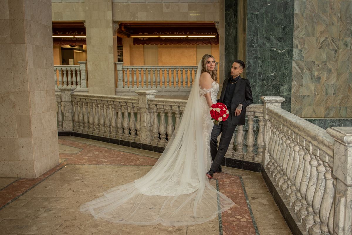 Bride in white gown and veil leans against groom in black suit near stone balustrade in grand atrium.