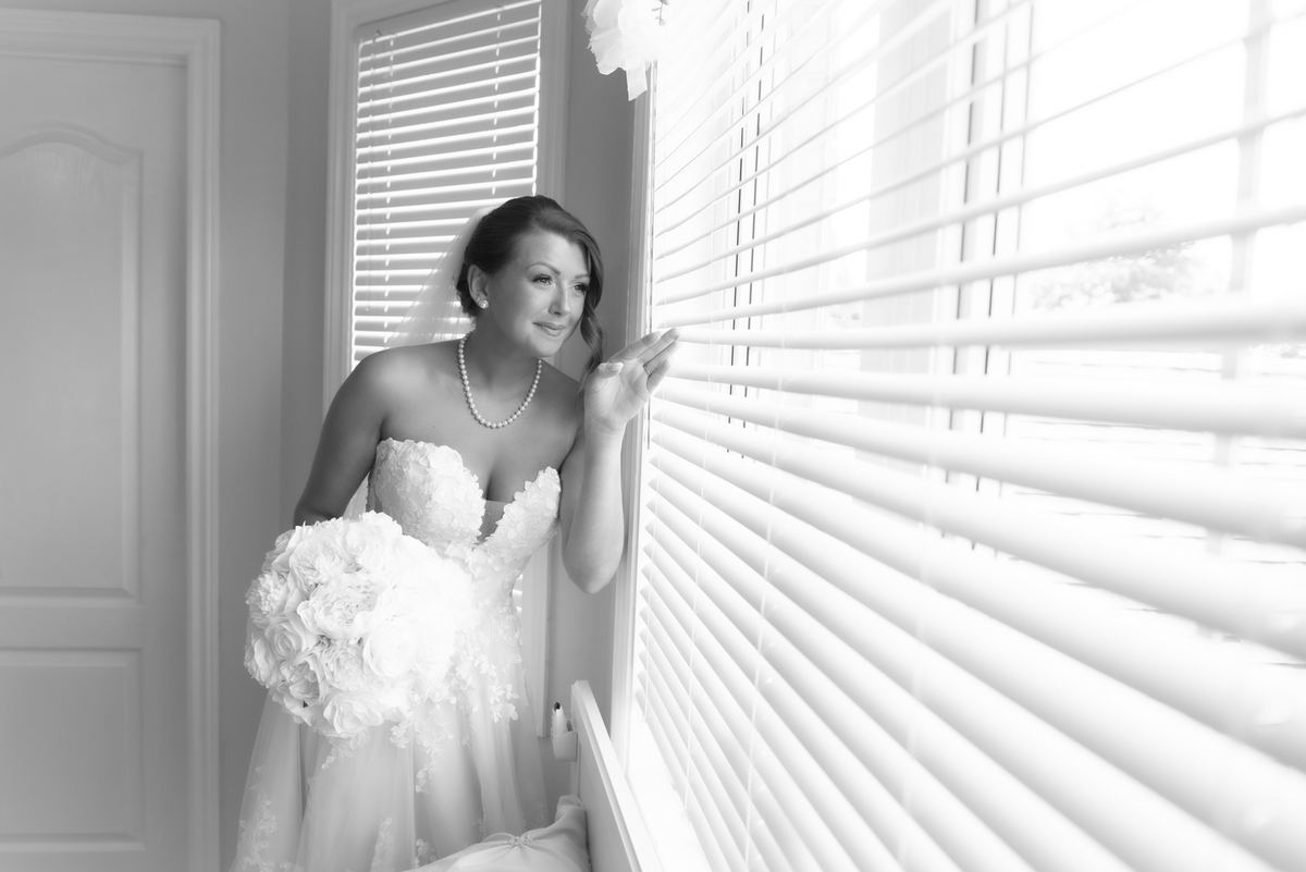 A bride in a white gown and veil stands by a window with horizontal blinds, holding a bouquet of white flowers.