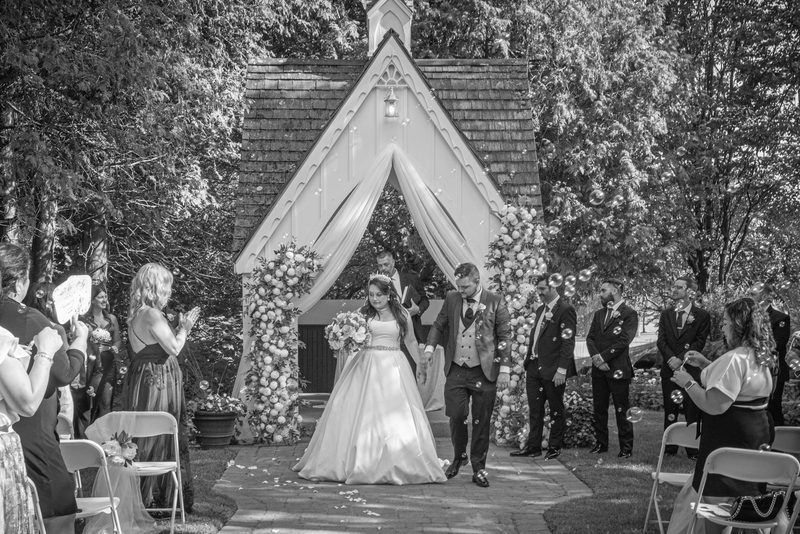 A bride and groom walk down the aisle under an arch, surrounded by applauding guests releasing bubbles.