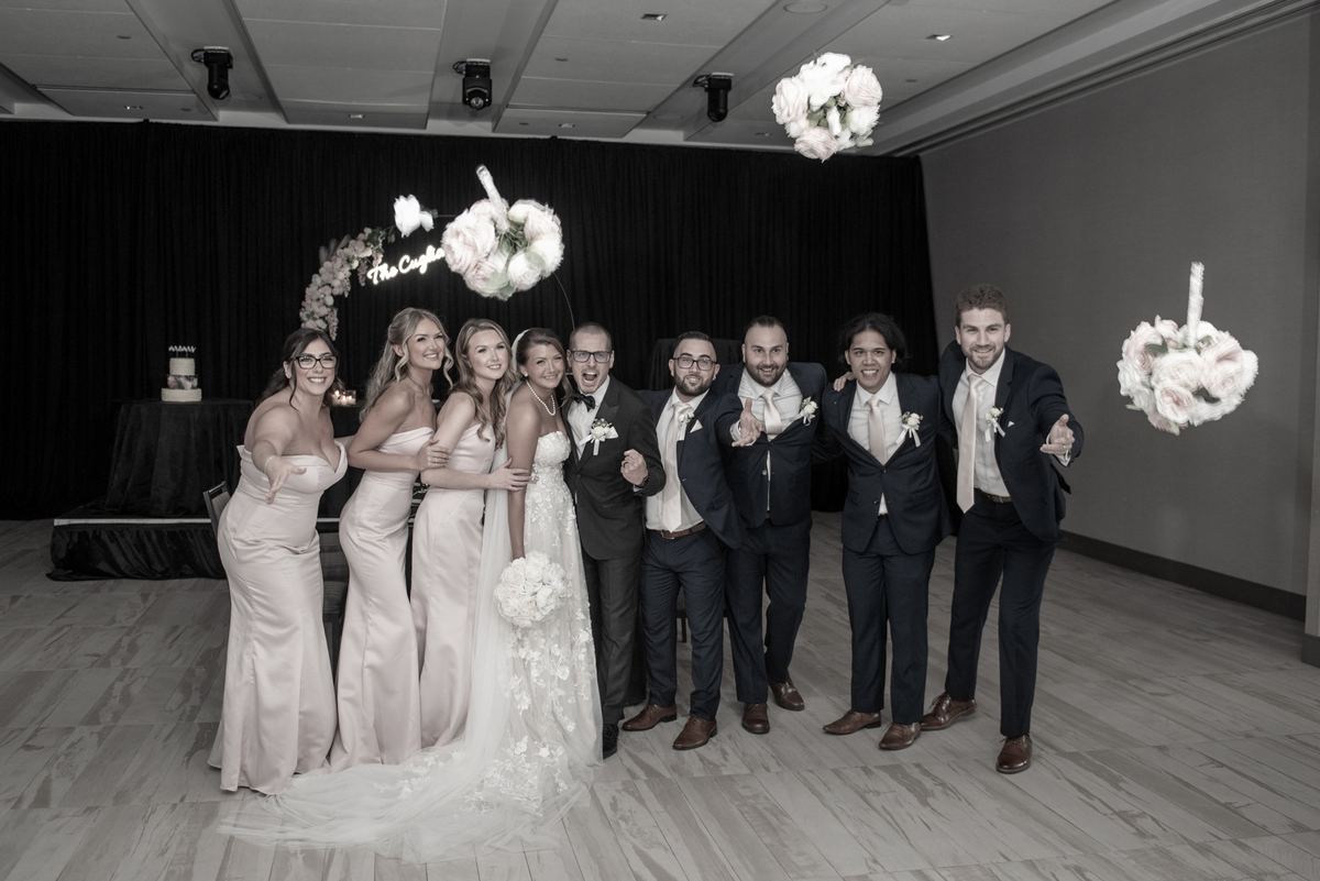 A bride and groom stand with their bridesmaids and groomsmen, all smiling amidst floating floral arrangements.