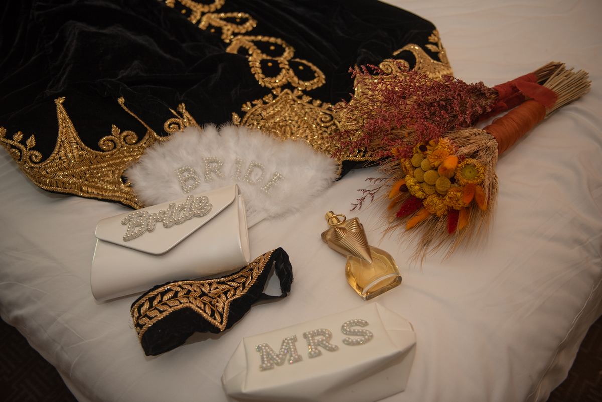 Bridal accessories including a robe, fan, clutch, headband, bouquet, and perfume bottle arranged on a bedspread.