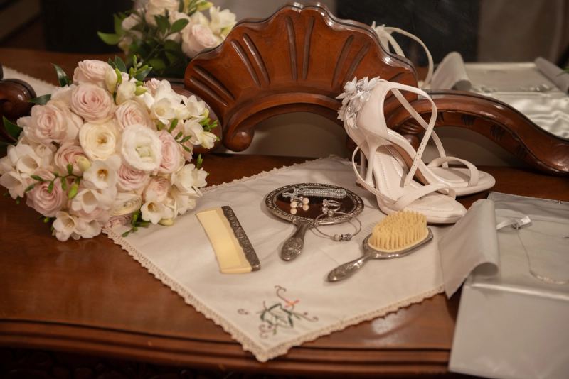 A polished wooden table set with white high-heeled shoes, pink and white flowers, silver grooming items.
