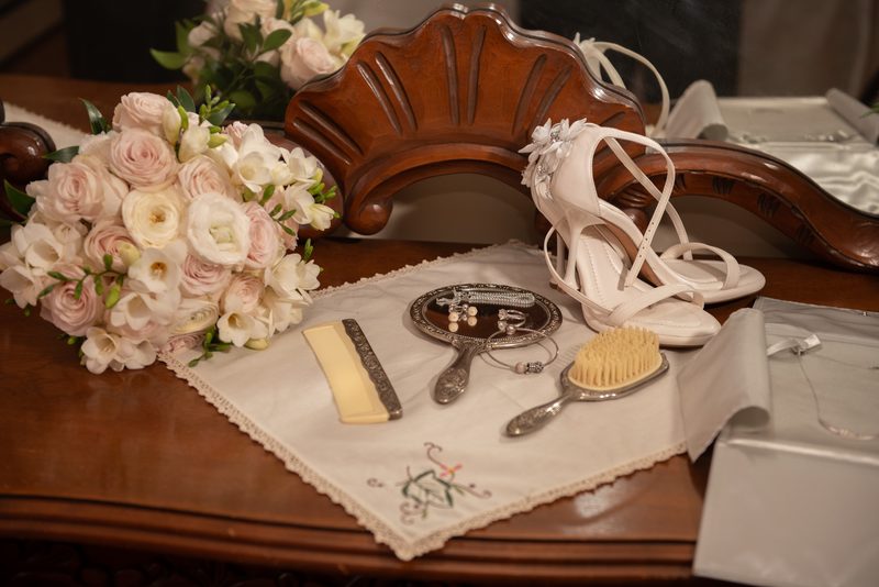 A polished wooden table set with white high-heeled shoes, pink and white flowers, silver grooming items.