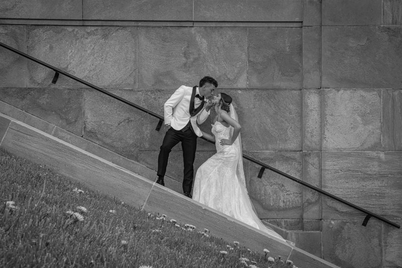 A bride and groom share a playful kiss leaning on a stone staircase railing above a grassy slope dotted with dandelions.