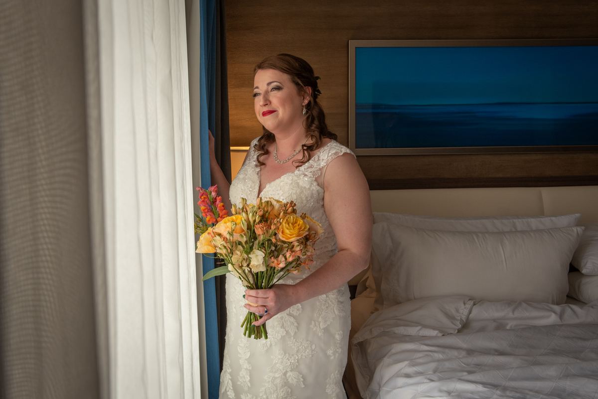 A bride holds orange and yellow flowers, peering out from behind a curtain in a hotel room bathed in soft light.