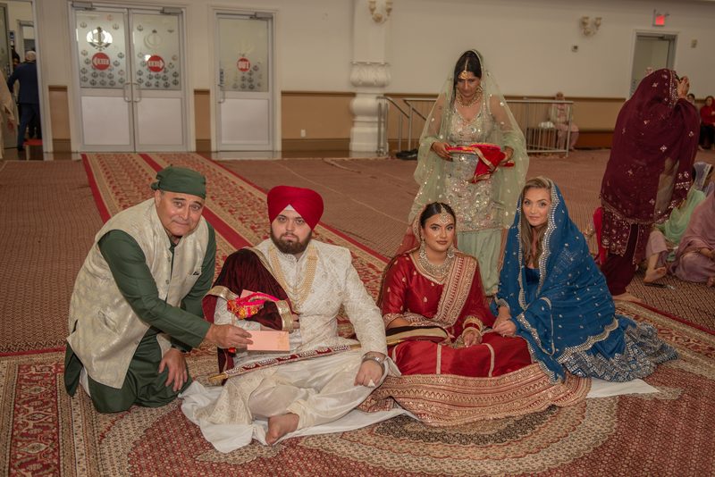 A Sikh bride and groom sit on a patterned carpet in a gurdwara, surrounded by family during a ceremony.