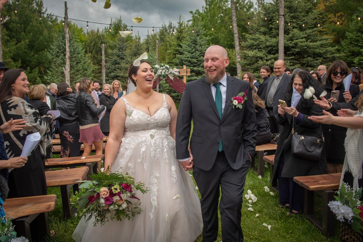 A smiling bride and groom walk down an outdoor aisle lined with guests throwing white petals, under pine trees.
