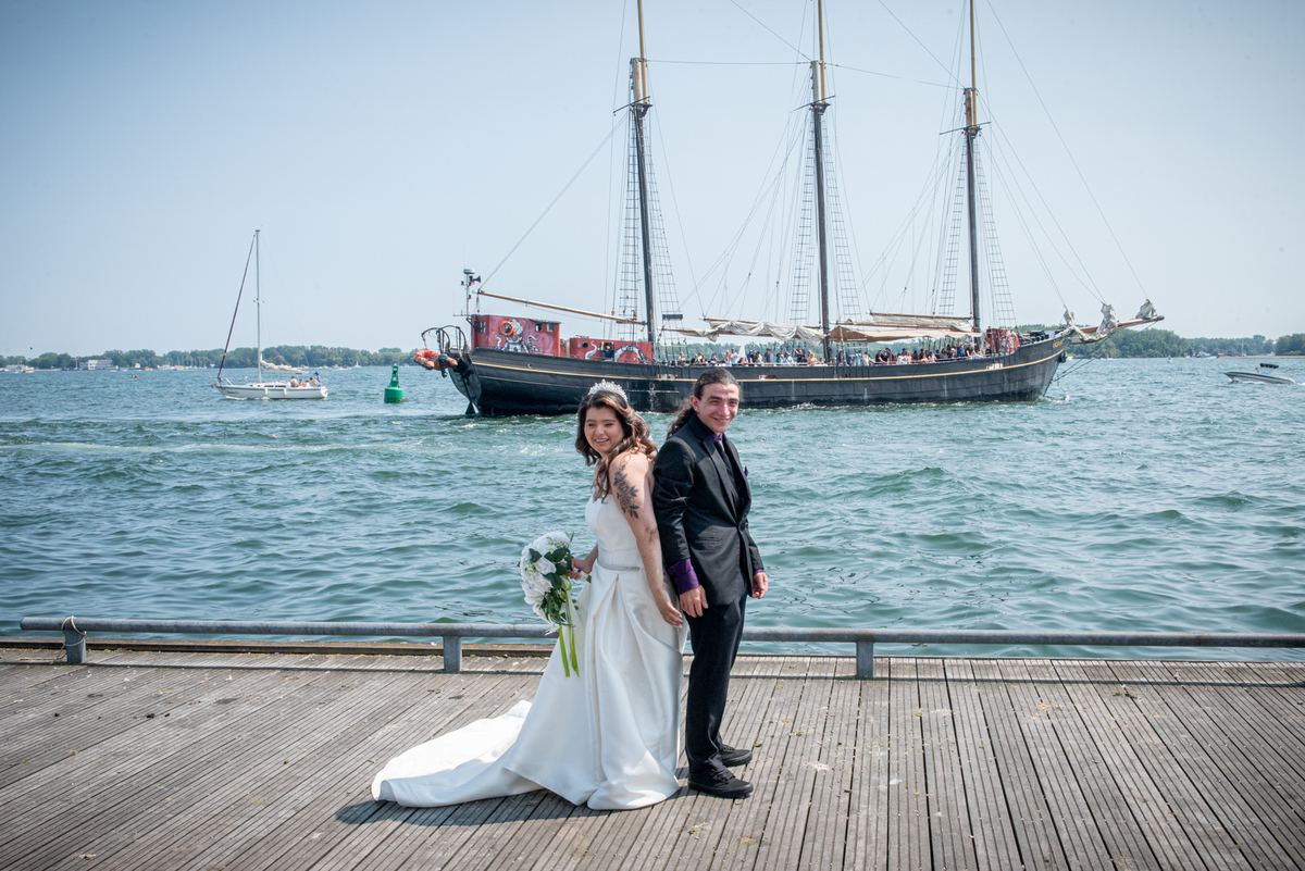 A bride and groom stand on a wooden pier, smiling with the historic sailing ship behind them in daylight.