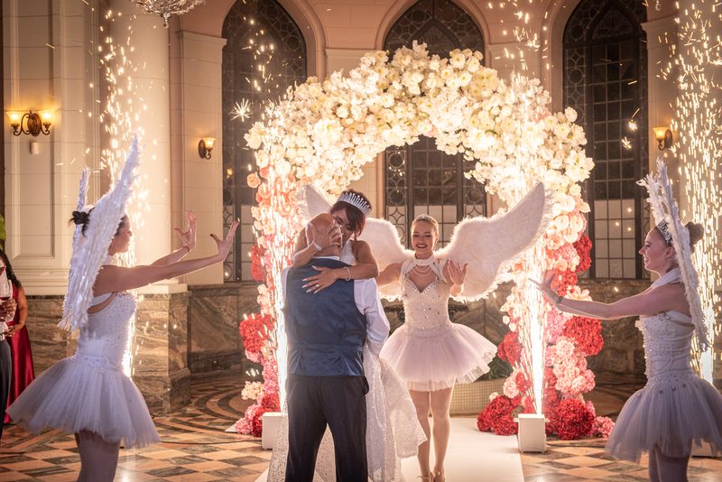 A bride and groom embrace under a floral arch, surrounded by angelic dancers in white tutos with wings, showering sparks.