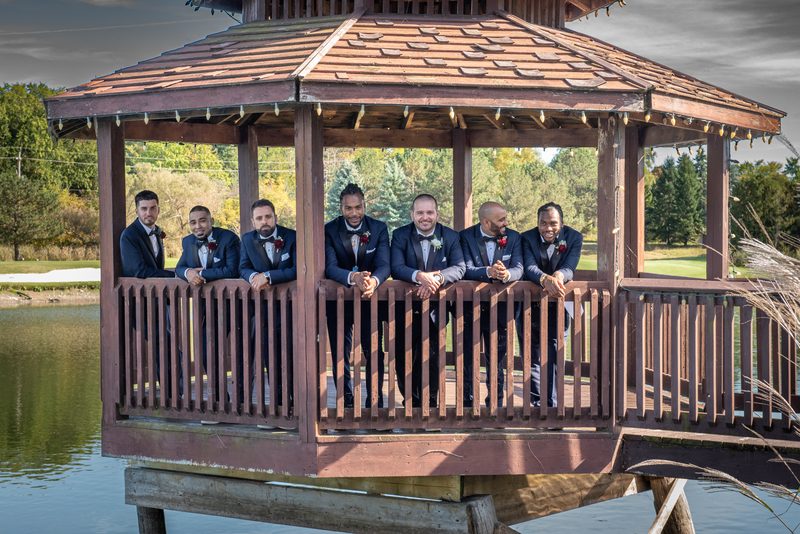 Seven groomsmen in tuxedos pose on a wooden gazebo overlooking water, smiling and leaning on the railing.