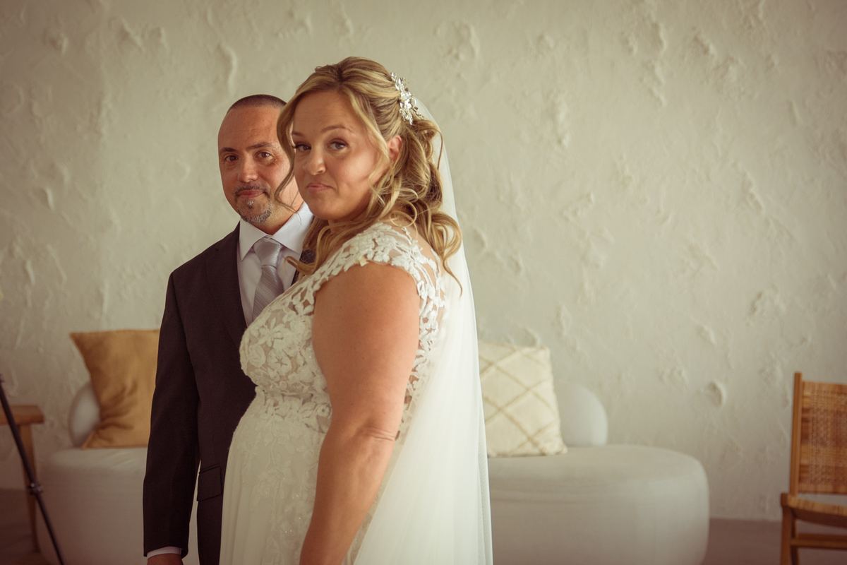 A bride turns to look back at the camera with her father standing behind her in a modern, minimalist indoor setting.