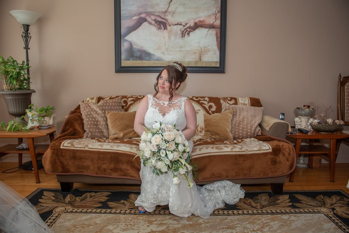 A bride in a white lace gown holds flowers and kneels with a smile in a living room decorated with a velvet sofa.